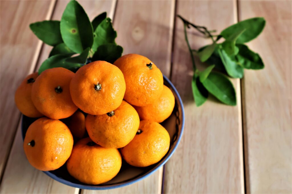 A bowl of clementines placed on a wooden table, with clementine leaves scattered beside the bowl and a few leaves resting on the bowl's corner.