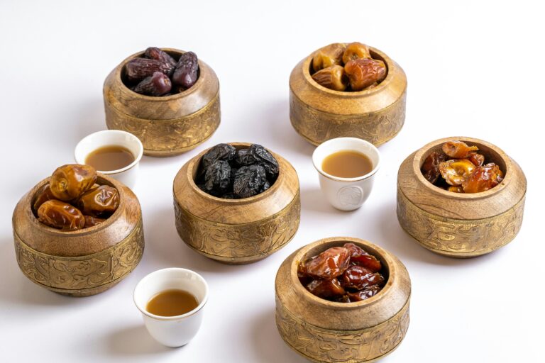 Six small wooden bowls filled with different types of dates, including one with Ajwa dates, and three white cups filled with a hot beverage, possibly Arabic tea or coffee, arranged on a surface.
