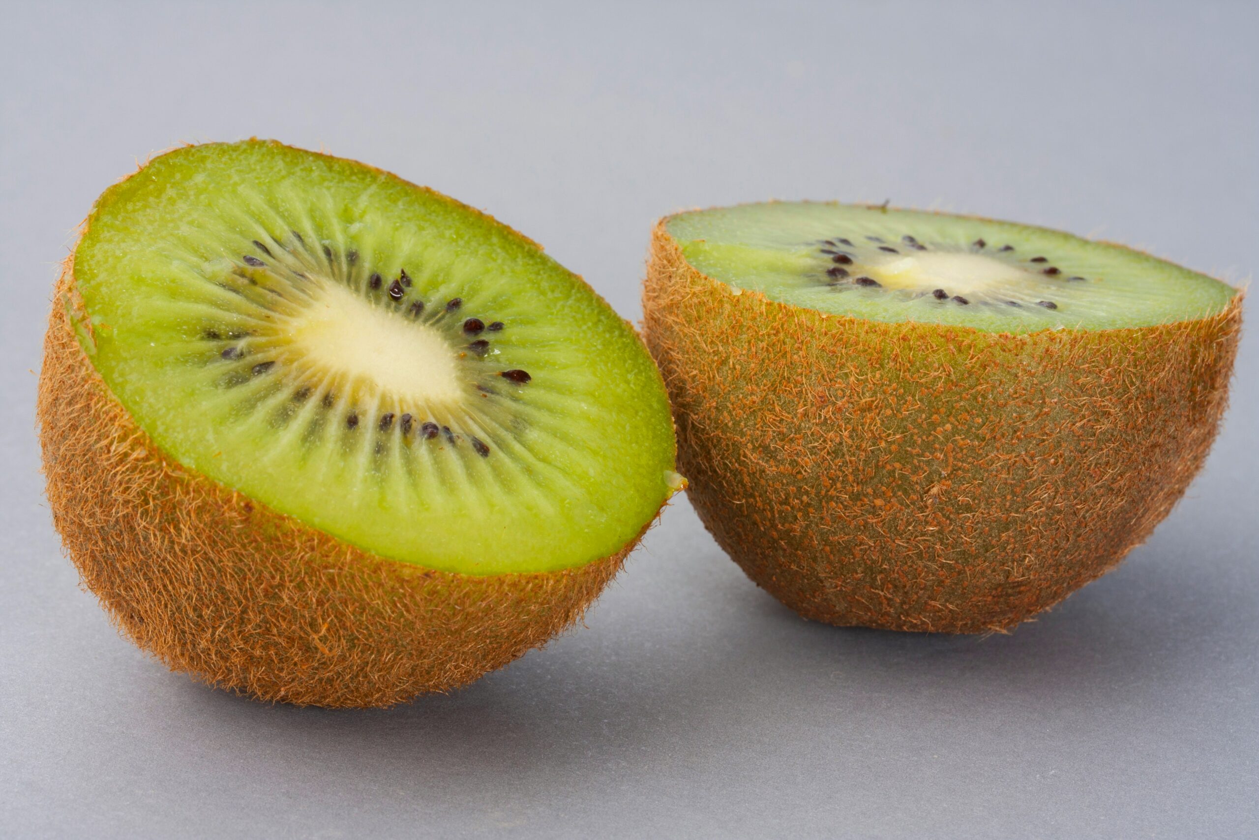 Two halves of a kiwi fruit placed on a grey background, cut cleanly to show the vibrant green flesh and black seeds.