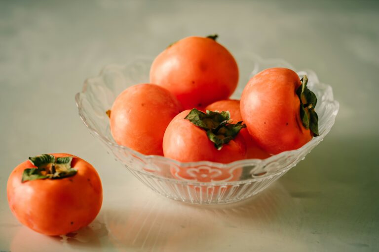 Five persimmons in a glass bowl and one persimmon placed beside the bowl on a wooden table.
