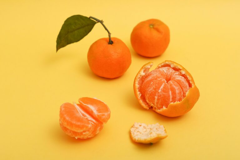 Two whole tangerines on a yellow background, one with a stitched-on leaf, the other half peeled with its skin lying beside it, along with one tangerine half placed nearby.